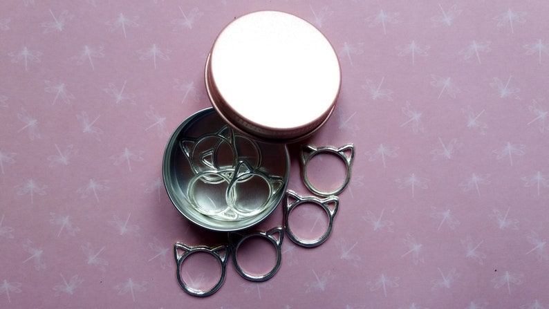 May include: A set of silver cat-shaped stitch markers in a round metal tin with a rose gold lid. The markers are arranged in the tin and some are scattered around it.
