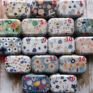 Pu&ograve; includere: Una collezione di sedici scatole di metallo rettangolari, ognuna con un design floreale o botanico unico. Le scatole presentano varie palette di colori, tra cui blu, verdi, arancioni e rosa, con motivi di fiori, foglie e forme astratte.