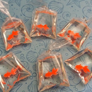 K&ouml;nnte beinhalten: Sechs transparente Plastikt&uuml;ten, jede mit einem leuchtend orangefarbenen Goldfisch. Die T&uuml;ten sind oben gebunden und auf einem hellblauen Hintergrund mit einem Muschelmuster angeordnet. Die Goldfische sind der Blickfang.