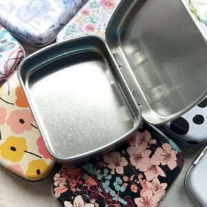K&ouml;nnte beinhalten: Eine offene, silberfarbene Metalldose liegt auf mehreren geschlossenen Dosen mit floralen und gemusterten Designs. Die geschlossenen Dosen weisen verschiedene Muster auf, darunter blaue Rosen, rosa Blumen und ein schwarzer Hintergrund mit wei&szlig;en Bl&uuml;ten.