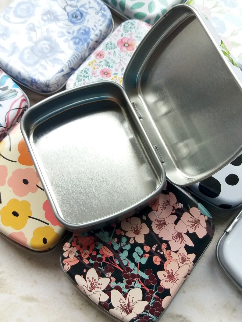 K&ouml;nnte beinhalten: Eine offene, silberfarbene Metalldose liegt auf mehreren geschlossenen Dosen mit floralen und gemusterten Designs. Die geschlossenen Dosen weisen verschiedene Muster auf, darunter blaue Rosen, rosa Blumen und ein schwarzer Hintergrund mit wei&szlig;en Bl&uuml;ten.