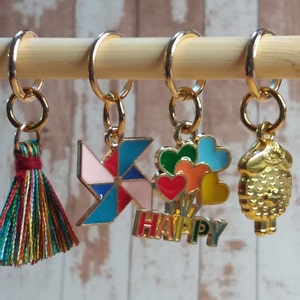 Pu&ograve; includere: Quattro marcapunti colorati con ciondoli diversi: un fiocco, una girandola rosa e blu, un mazzo di cuori colorati con la scritta "HAPPY" sotto e una pecora dorata. I marcapunti sono appesi a un bastoncino di legno.