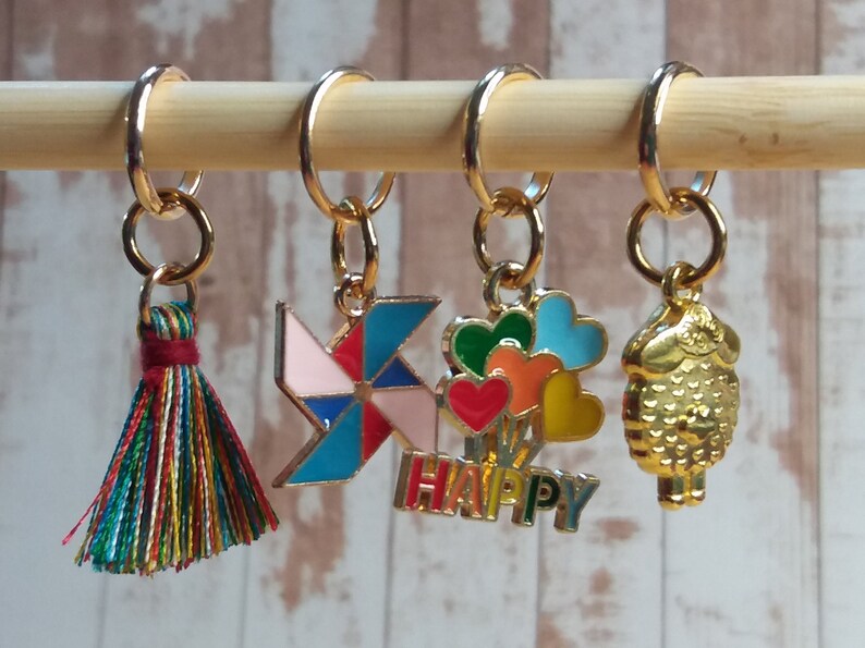 Pu&ograve; includere: Quattro marcapunti colorati con ciondoli diversi: un fiocco, una girandola rosa e blu, un mazzo di cuori colorati con la scritta "HAPPY" sotto e una pecora dorata. I marcapunti sono appesi a un bastoncino di legno.
