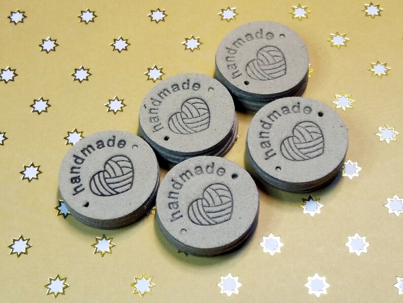 May include: Five brown round paper tags with the word "handmade" and a heart-shaped yarn ball design. The tags are on a gold background with white stars.