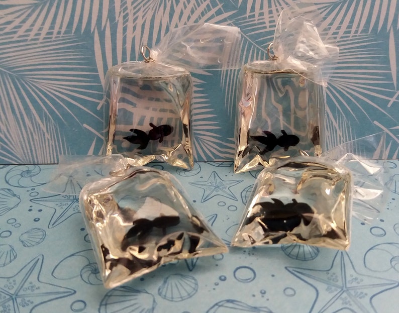 K&ouml;nnte beinhalten: Vier transparente, beutelf&ouml;rmige Ornamente, die jeweils einen schwarzen Fisch enthalten. Die Beutel sind oben mit einer goldfarbenen Schlaufe zum Aufh&auml;ngen verschlossen. Die Ornamente sind auf einer blau-wei&szlig; gemusterten Oberfl&auml;che angeordnet.