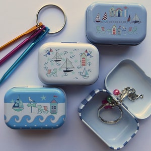 Op de afbeelding: Vier kleine blauwe en witte metalen blikken met nautische ontwerpen. De blikken zijn versierd met zeilboten, strandhuisjes en zeemeeuwen. Een blik is open en bevat een zilveren sleutelhanger met een bedel.