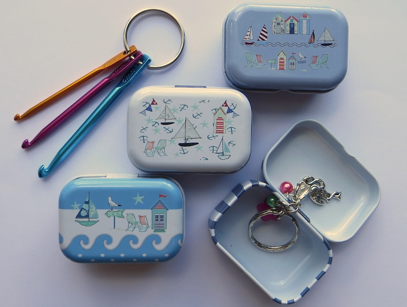 Op de afbeelding: Vier kleine blauwe en witte metalen blikken met nautische ontwerpen. De blikken zijn versierd met zeilboten, strandhuisjes en zeemeeuwen. Een blik is open en bevat een zilveren sleutelhanger met een bedel.
