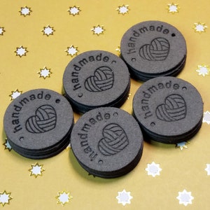 May include: Five round, black, leather tags with the word "handmade" and a heart with yarn inside. The tags have a hole punched in the top.