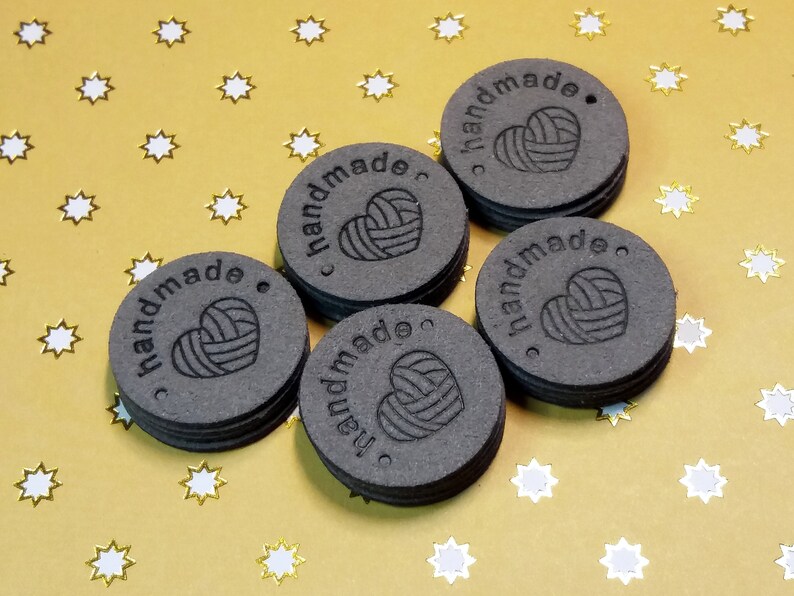 May include: Five round, black, leather tags with the word "handmade" and a heart with yarn inside. The tags have a hole punched in the top.