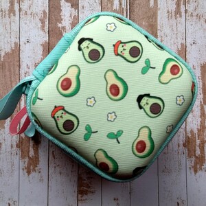 Puede incluir: Estuche cuadrado con cremallera, con un fondo verde claro y un estampado de aguacates. Los aguacates tienen caras y llevan sombreros. El estuche tiene una cremallera turquesa y un lazo turquesa con un acento rosa.