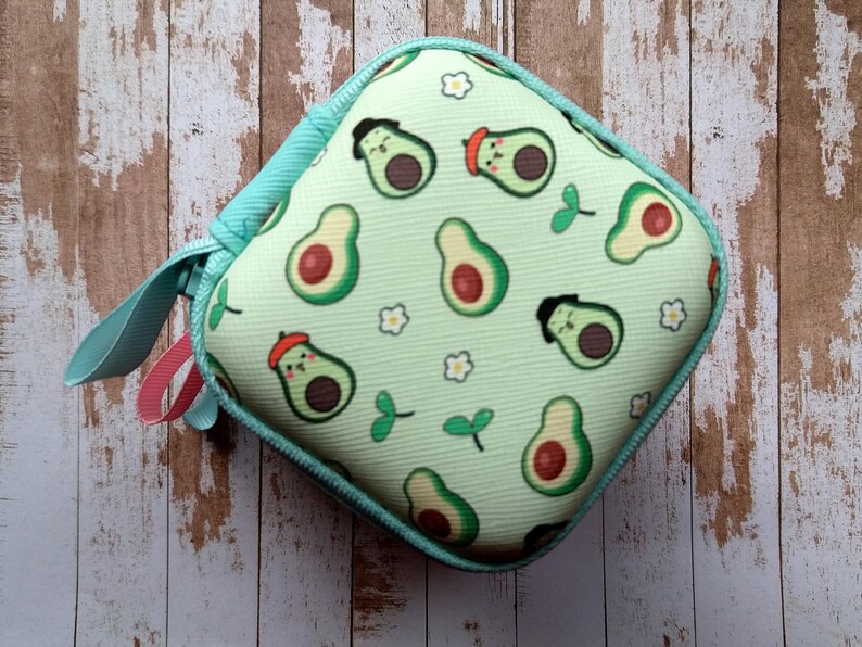 Puede incluir: Estuche cuadrado con cremallera, con un fondo verde claro y un estampado de aguacates. Los aguacates tienen caras y llevan sombreros. El estuche tiene una cremallera turquesa y un lazo turquesa con un acento rosa.