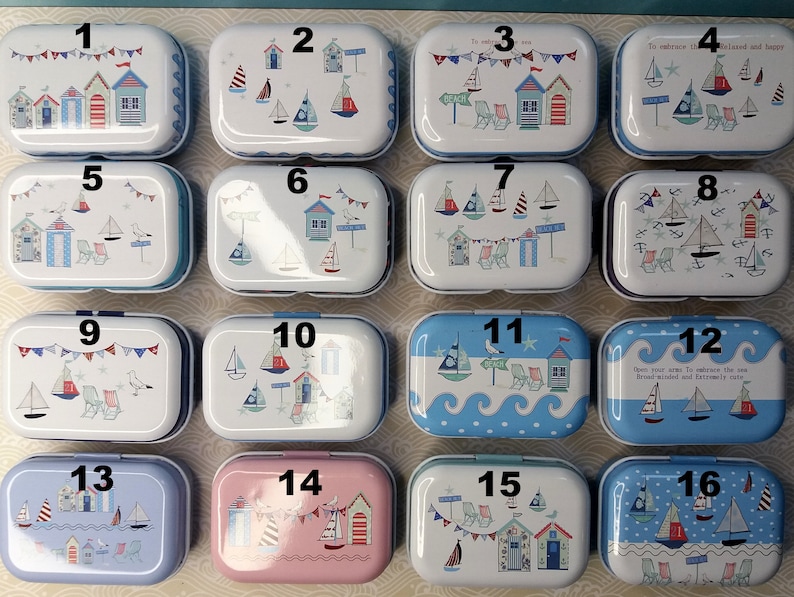 Op de afbeelding: Set van 16 kleine, rechthoekige, metalen blikken met verschillende nautische ontwerpen. Elk blik heeft een nummer van 1 tot 16 erop gedrukt. De blikken zijn wit, blauw, roze en lichtblauw met afbeeldingen van zeilboten, strandhuisjes en schelpen.