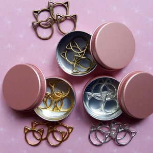 May include: Three pink metal containers with lids, each containing a set of gold or silver cat-shaped ring holders. The containers are arranged in a triangle shape on a pink background with a white pattern.