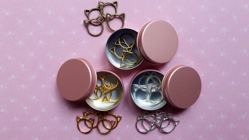 May include: Three pink metal containers with lids, each containing a set of gold or silver cat-shaped ring holders. The containers are arranged in a triangle shape on a pink background with a white pattern.