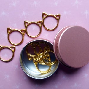May include: A set of gold cat-shaped stitch markers in a pink metal container. The markers are arranged in a line on a pink background with a white pattern.