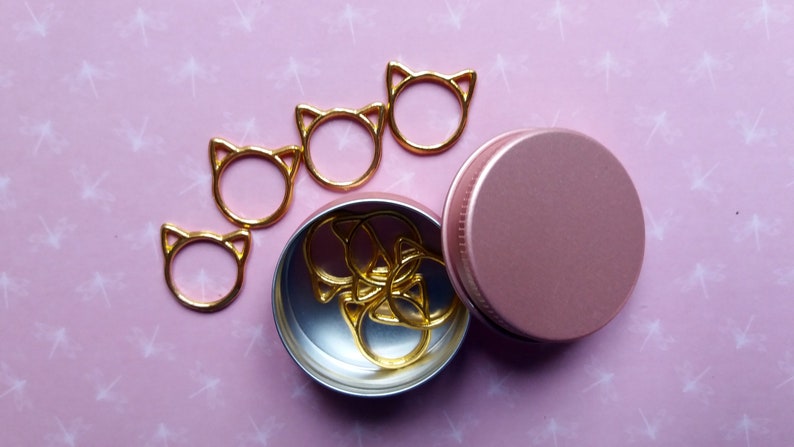 May include: A set of gold cat-shaped stitch markers in a pink metal container. The markers are arranged in a line on a pink background with a white pattern.