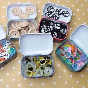 Pu&ograve; includere: Una collezione di piccole scatole di metallo aperte piene di vari materiali per l'artigianato. Le scatole contengono bottoni di legno, perline con numeri, segnapunti colorati e altri abbellimenti. Lo sfondo &egrave; una superficie gialla con pois bianchi.