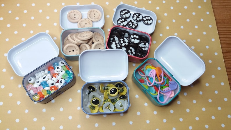 Pu&ograve; includere: Una collezione di piccole scatole di metallo aperte piene di vari materiali per l'artigianato. Le scatole contengono bottoni di legno, perline con numeri, segnapunti colorati e altri abbellimenti. Lo sfondo &egrave; una superficie gialla con pois bianchi.