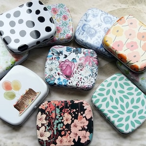 K&ouml;nnte beinhalten: Eine Sammlung dekorativer rechteckiger Dosen mit verschiedenen floralen, gepunkteten und grafischen Designs. Die Dosen weisen eine Vielzahl von Farben auf, darunter Rosa, Blau, Wei&szlig; und Schwarz. Einige Dosen haben Bilder von Kuchen und Luftballons.