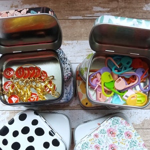 K&ouml;nnte beinhalten: Vier kleine, offene Metalldosen mit dekorativen Deckeln. Zwei Dosen enthalten Strickzubeh&ouml;r: rote Maschenmarkierer und bunte Maschenmarkierer aus Kunststoff. Die anderen beiden Dosen haben schwarze und wei&szlig;e Tupfen und ein Blumenmuster.
