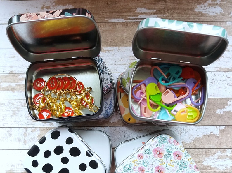 K&ouml;nnte beinhalten: Vier kleine, offene Metalldosen mit dekorativen Deckeln. Zwei Dosen enthalten Strickzubeh&ouml;r: rote Maschenmarkierer und bunte Maschenmarkierer aus Kunststoff. Die anderen beiden Dosen haben schwarze und wei&szlig;e Tupfen und ein Blumenmuster.