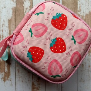 Puede incluir: Un estuche cuadrado rosa con cremallera y estampado de fresas. El dise&ntilde;o presenta fresas rojas con tallos verdes y mitades de fresas rosas, junto con texto japon&eacute;s. Un lazo rosa est&aacute; unido a la cremallera.