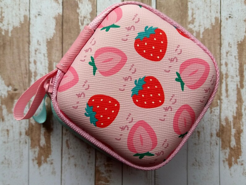 Puede incluir: Un estuche cuadrado rosa con cremallera y estampado de fresas. El dise&ntilde;o presenta fresas rojas con tallos verdes y mitades de fresas rosas, junto con texto japon&eacute;s. Un lazo rosa est&aacute; unido a la cremallera.