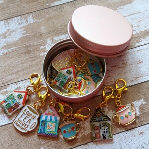 May include: A pink metal container with a lid, filled with colorful stitch markers with various designs, including houses, teacups, and a mug that says "A Cup of Tea Solves Everything".