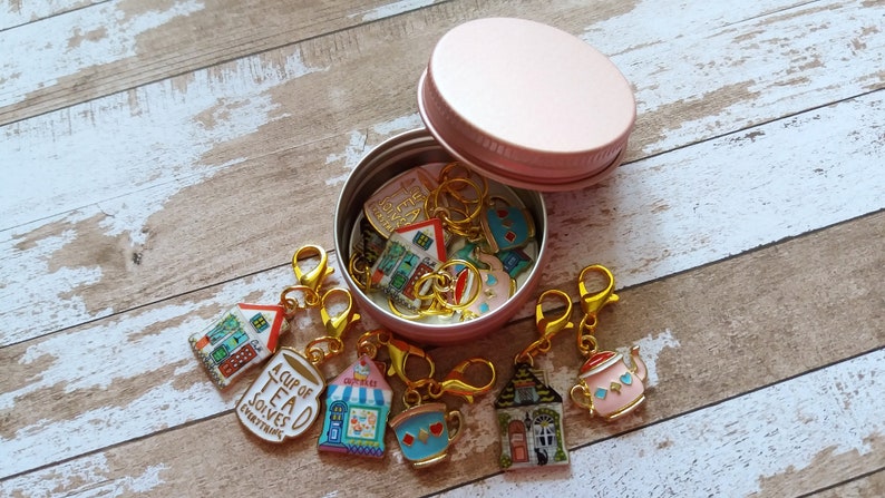 May include: A pink metal container with a lid, filled with colorful stitch markers with various designs, including houses, teacups, and a mug that says "A Cup of Tea Solves Everything".