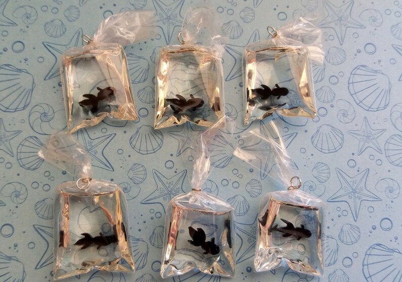 K&ouml;nnte beinhalten: Sechs klare Plastikt&uuml;ten, jede mit einem kleinen schwarzen Fisch, sind auf einem hellblauen Hintergrund mit einem Muschelmuster angeordnet. Die T&uuml;ten sind oben mit einem silbernen Ring zum Aufh&auml;ngen gebunden.