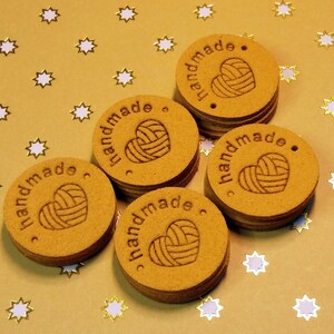 May include: Six round, brown, wooden tags with the word "handmade" and a heart-shaped yarn ball design. The tags are on a gold background with white stars.