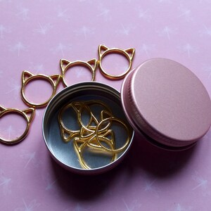 May include: A set of gold cat-shaped stitch markers in a pink metal container. The container has a lid and is open, revealing the markers inside.