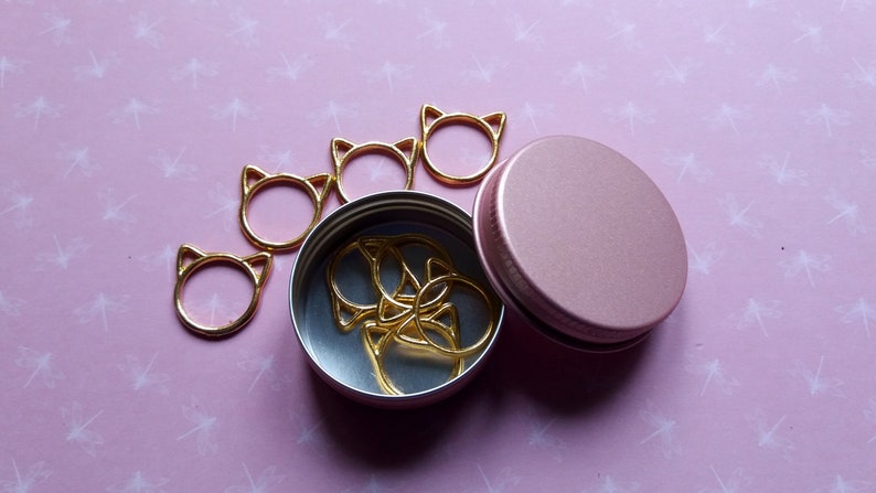 May include: A set of gold cat-shaped stitch markers in a pink metal container. The container has a lid and is open, revealing the markers inside.