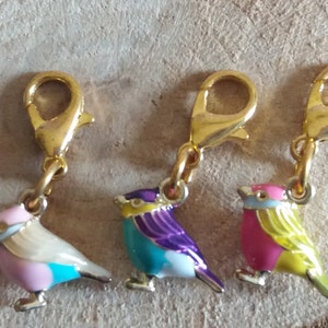 May include: Three gold bird charms with enamel paint. The birds are pink and yellow, blue and purple, and white and pink.
