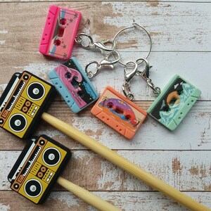 Könnte beinhalten: Eine Sammlung bunter Kassettenband-Anhänger und zwei gelb-schwarze Boombox-Anhänger. Die Kassettenband-Anhänger sind rosa, blau, orange und grün, jeweils mit einzigartigen Designs. Silberne Verschlüsse und ein Schlüsselring sind angebracht.