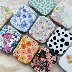 K&ouml;nnte beinhalten: Eine Sammlung kleiner, rechteckiger Metalldosen mit verschiedenen floralen und gemusterten Designs. Die Dosen zeigen Designs wie blaue Rosen, rosa Blumen und ein schwarz-wei&szlig;es Tupfenmuster. Einige Dosen haben den Text "Love Never Ends".