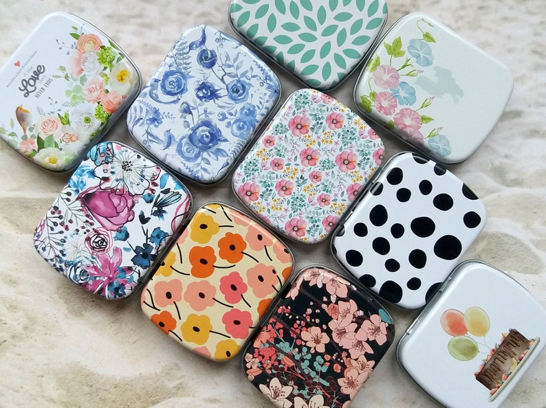 K&ouml;nnte beinhalten: Eine Sammlung kleiner, rechteckiger Metalldosen mit verschiedenen floralen und gemusterten Designs. Die Dosen zeigen Designs wie blaue Rosen, rosa Blumen und ein schwarz-wei&szlig;es Tupfenmuster. Einige Dosen haben den Text "Love Never Ends".