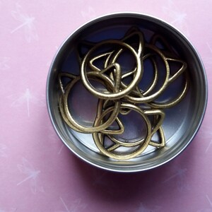 May include: A small metal container filled with gold cat-shaped rings. The container is round and silver.