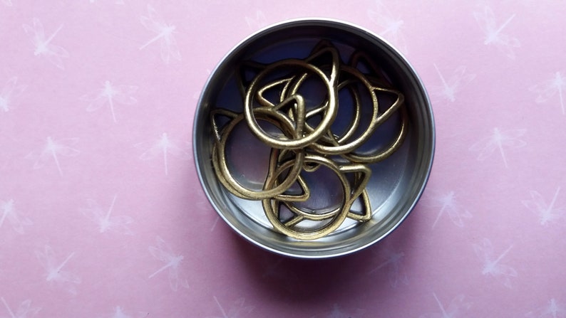 May include: A small metal container filled with gold cat-shaped rings. The container is round and silver.