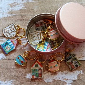 May include: A set of 10 gold-toned stitch markers with teacup and teapot designs. The markers are in a pink metal tin with the words "A Cup of Tea Solves Everything".