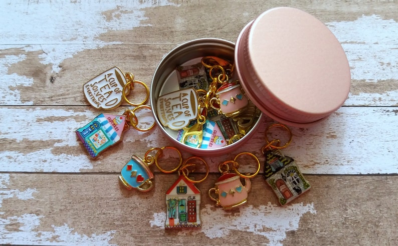 May include: A set of 10 gold-toned stitch markers with teacup and teapot designs. The markers are in a pink metal tin with the words "A Cup of Tea Solves Everything".