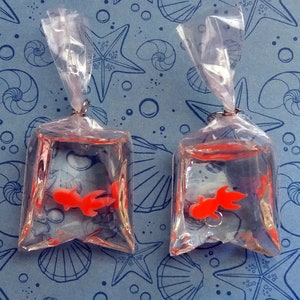 K&ouml;nnte beinhalten: Zwei transparente Plastikt&uuml;ten, jede mit einem kleinen orangefarbenen Goldfisch, sind an silbernen Reifen aufgeh&auml;ngt. Die T&uuml;ten sind mit klarer Fl&uuml;ssigkeit gef&uuml;llt, und die Goldfische sind darin sichtbar. Der Hintergrund ist ein blauer Stoff mit einem Muschel- und Seesternmuster.