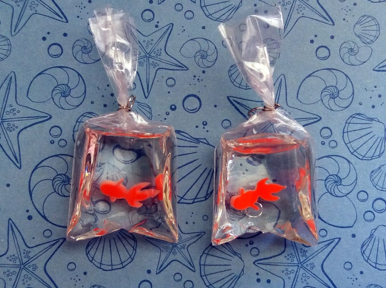 K&ouml;nnte beinhalten: Zwei transparente Plastikt&uuml;ten, jede mit einem kleinen orangefarbenen Goldfisch, sind an silbernen Reifen aufgeh&auml;ngt. Die T&uuml;ten sind mit klarer Fl&uuml;ssigkeit gef&uuml;llt, und die Goldfische sind darin sichtbar. Der Hintergrund ist ein blauer Stoff mit einem Muschel- und Seesternmuster.
