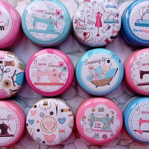 Op de afbeelding: Een verzameling ronde, decoratieve blikken met naaigerelateerde ontwerpen. De blikken tonen afbeeldingen van naaimachines, garenklosjes, scharen en de tekst "Tailor Shop", "I love Sewing" en "Sewing Time". De blikken zijn in tinten roze, blauw en wit.