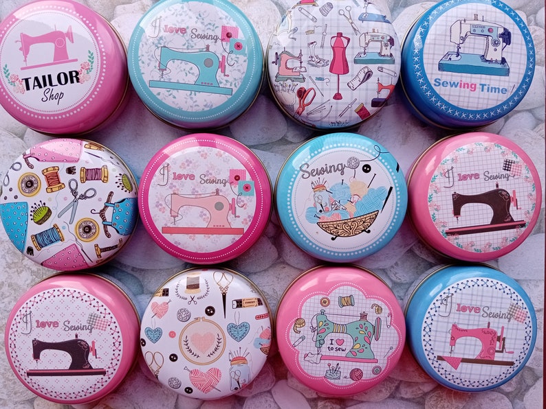 Op de afbeelding: Een verzameling ronde, decoratieve blikken met naaigerelateerde ontwerpen. De blikken tonen afbeeldingen van naaimachines, garenklosjes, scharen en de tekst "Tailor Shop", "I love Sewing" en "Sewing Time". De blikken zijn in tinten roze, blauw en wit.