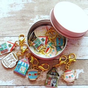 May include: A set of 10 colorful enamel charms in a pink tin. The charms feature teacups, teapots, and houses with whimsical designs. The charms are attached to gold-colored lobster clasps.