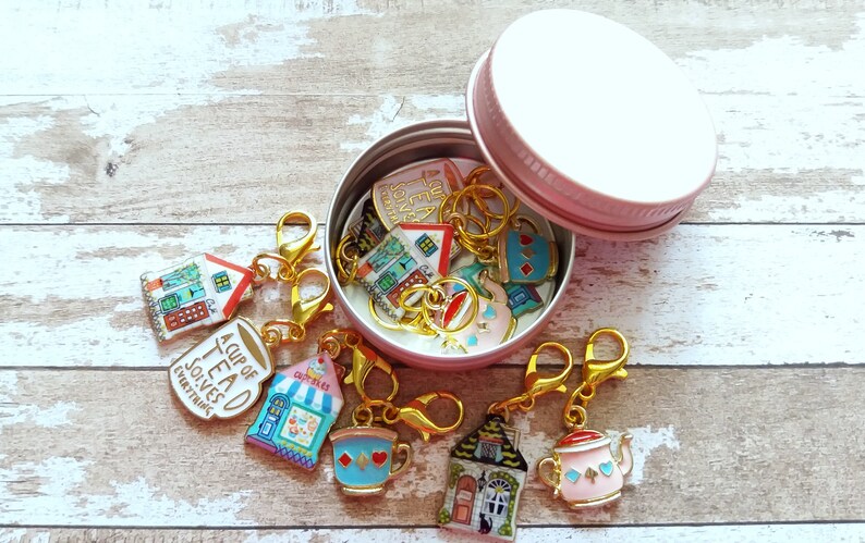 May include: A set of 10 colorful enamel charms in a pink tin. The charms feature teacups, teapots, and houses with whimsical designs. The charms are attached to gold-colored lobster clasps.
