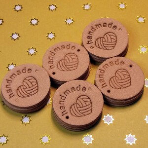 May include: Five brown leather tags with the word "handmade" and a heart with yarn inside. The tags are on a gold background with white stars.