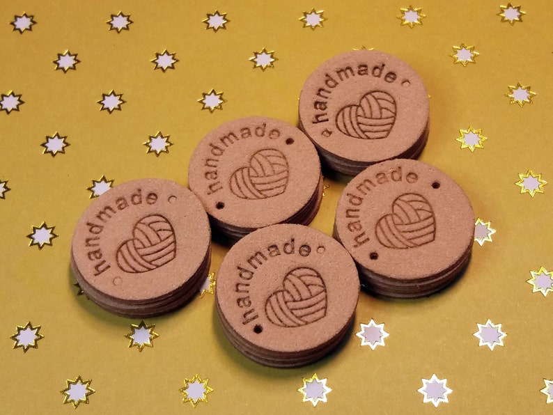 May include: Five brown leather tags with the word "handmade" and a heart with yarn inside. The tags are on a gold background with white stars.