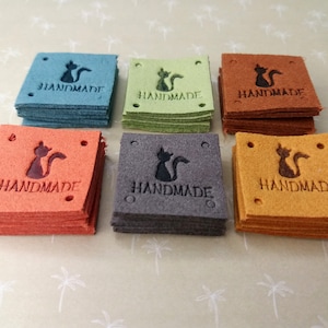 Peut inclure: Six piles de tags en cuir avec une silhouette de chat et le mot "HANDMADE" en lettres noires. Les étiquettes sont de six couleurs différentes : sarcelle, vert, marron, orange, gris et jaune.